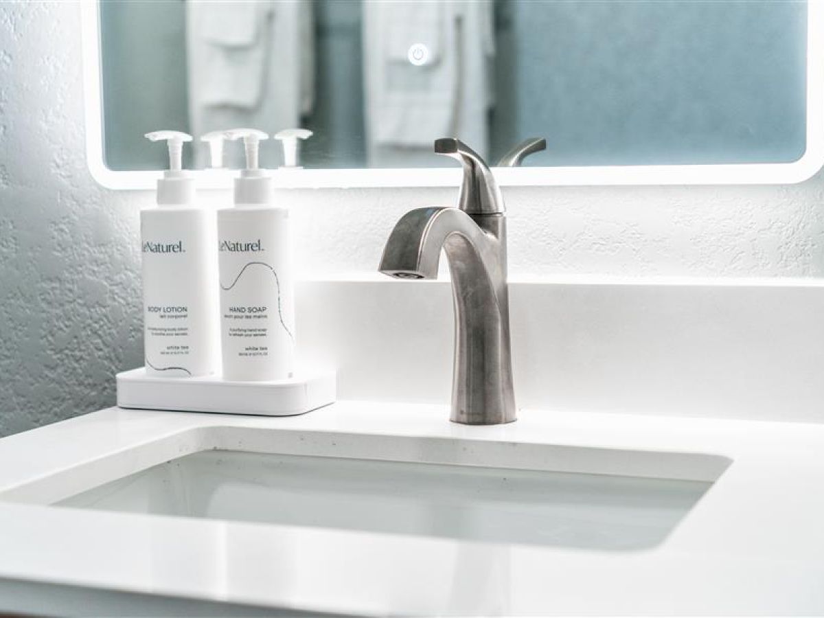 A modern bathroom sink with a sleek silver faucet and two bottles of hand care products on the countertop, next to a backlit mirror.