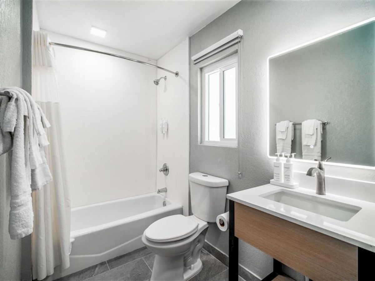 A modern bathroom featuring a bathtub, toilet, sink with a backlit mirror, towels, and soap dispensers, all in a neutral color scheme.