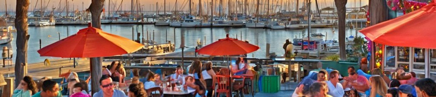 People dining outdoors at a waterfront restaurant with orange umbrellas, palm trees, and sailboats in the background, enjoying a warm sunset.